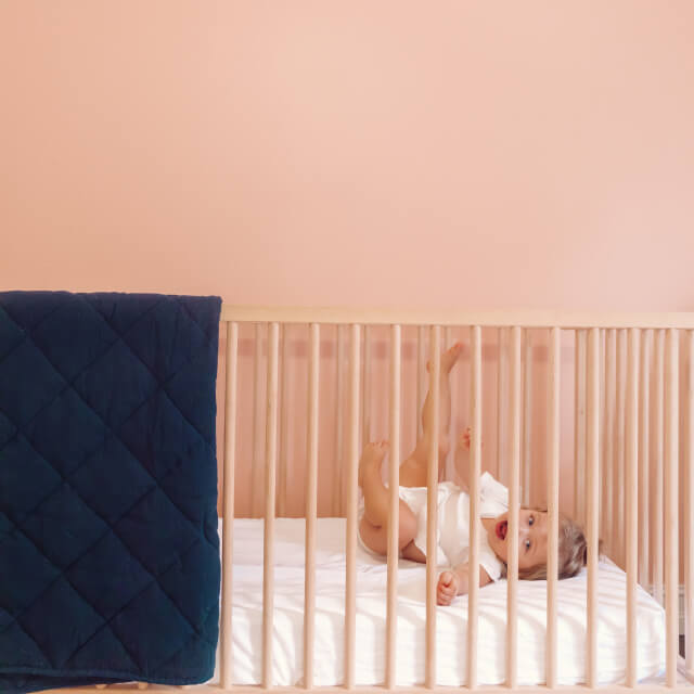 A young child laughing and lying in a traditional crib with the Slumber Cloud Essential Fitted Sheet - Crib in white