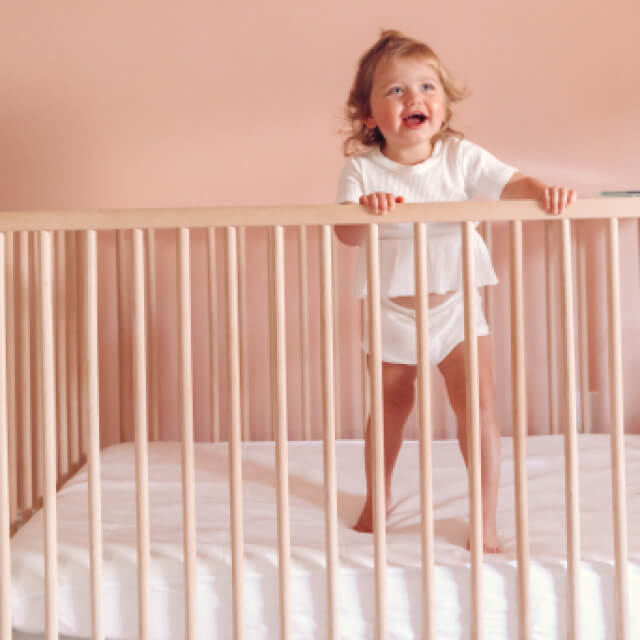 A young child laughing and standing in a traditional crib with the Slumber Cloud Essential Fitted Sheet - Crib in white