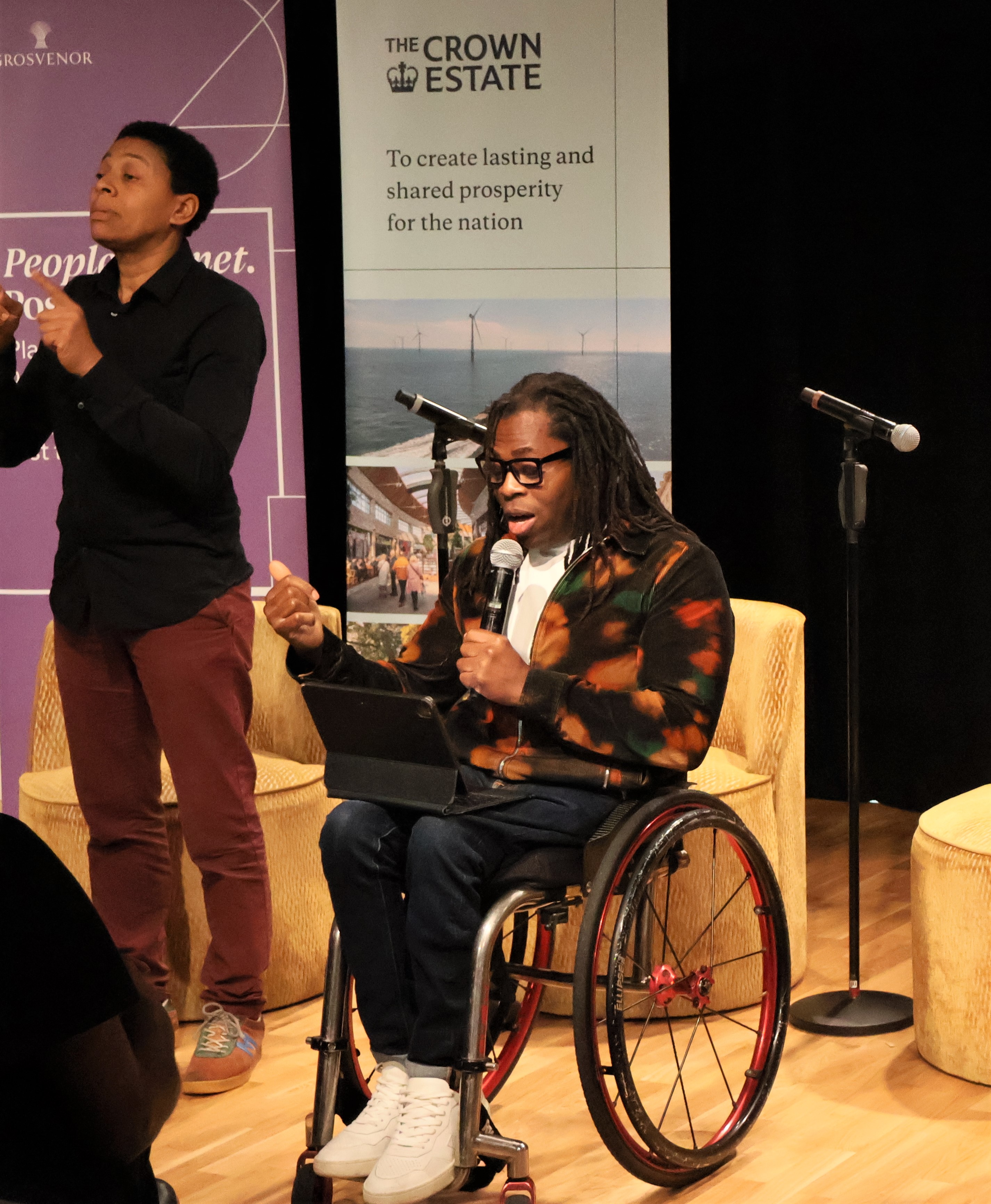 Ade Adepitan delivering keynote speech on stage at Inclusive Spaces and Places initiative launch beside BSL translator