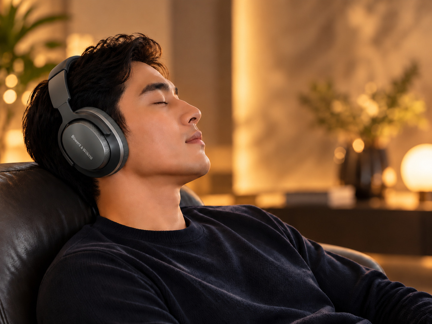 Young man relaxing indoors while wearing Bowers and Wilkins over-ear wireless headphones in warm ambient lighting