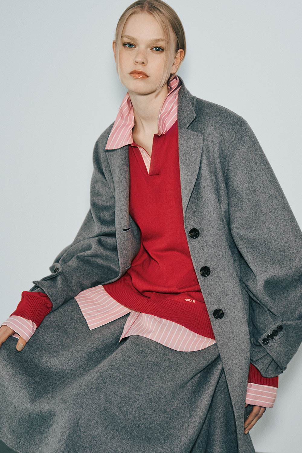 The model wears an oversized gray wool coat with black buttons over a red V-neck knit sweater and a pink-and-white striped shirt, paired with a gray skirt.