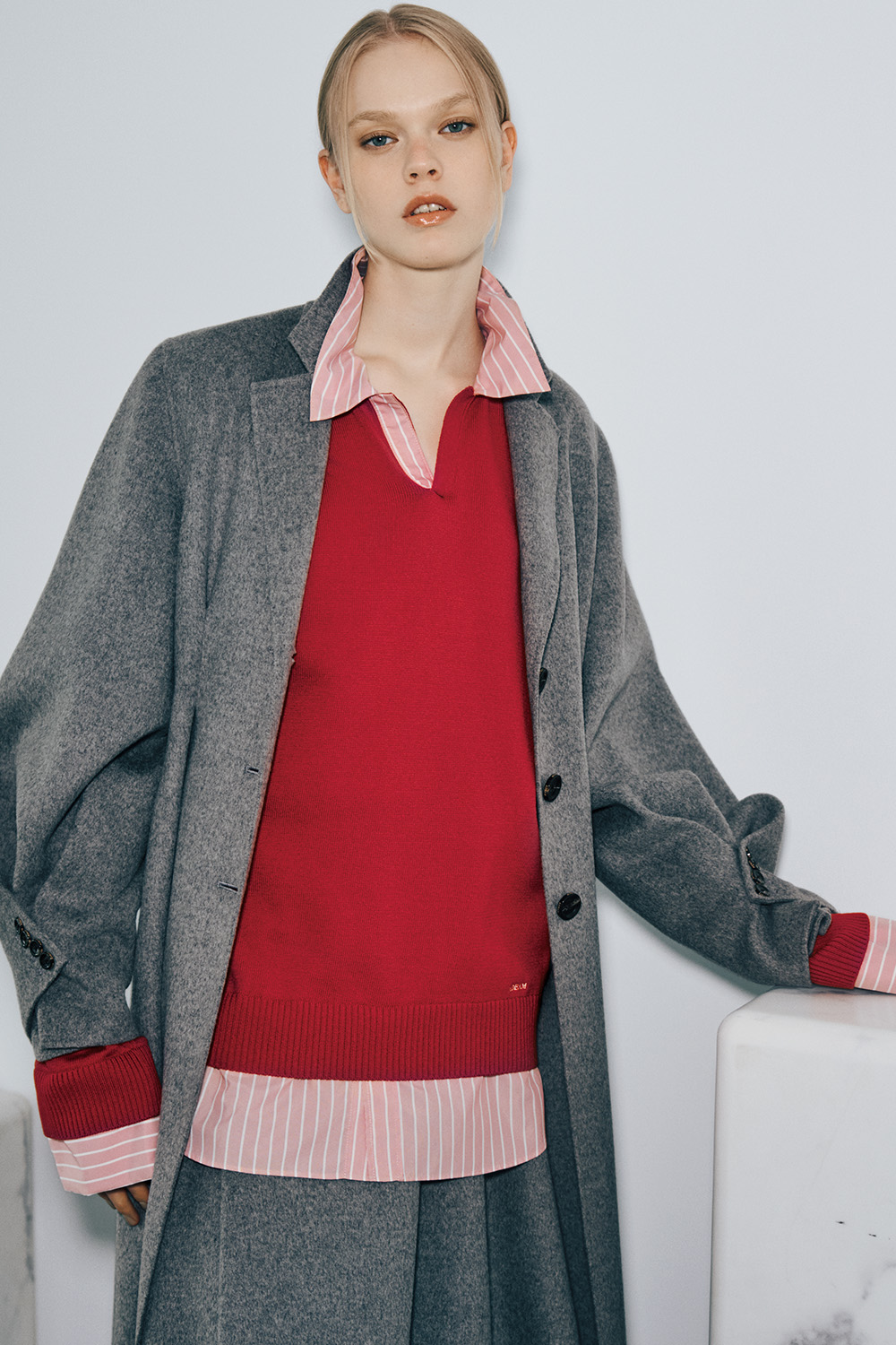 The model wears an oversized gray wool coat open over a red V-neck knit sweater and a pink-and-white striped shirt, with the shirt collar, cuffs, and hem showing, and a gray skirt.