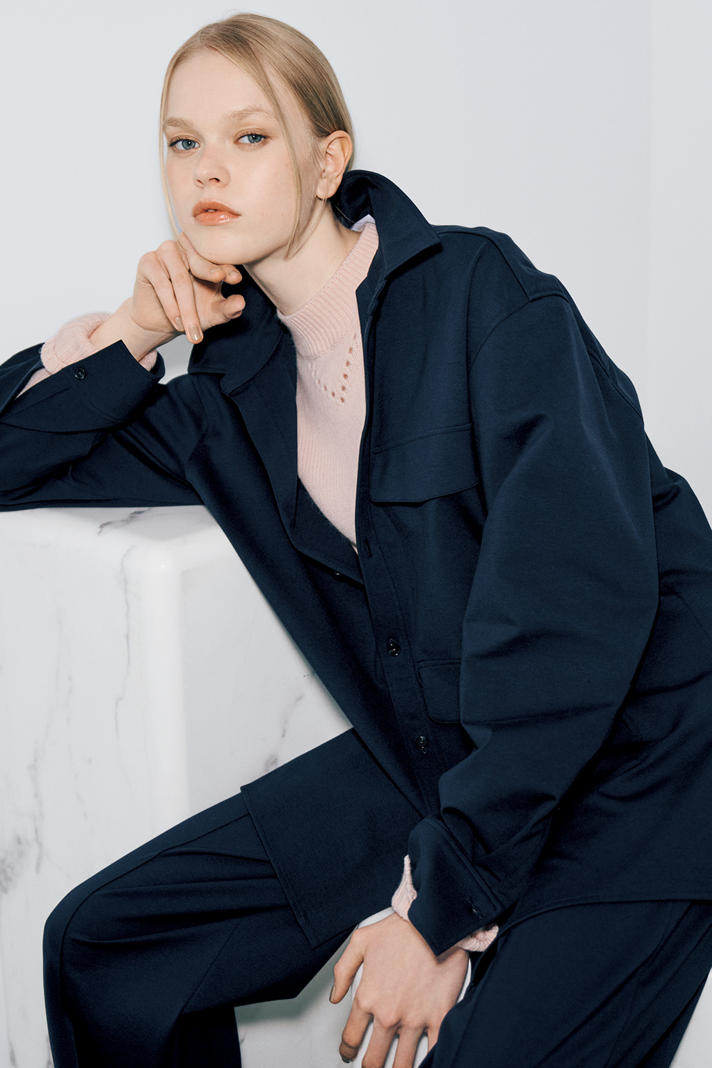 The model wears a navy oversized shirt jacket over a pale pink turtleneck with openwork at the neckline, and navy trousers.