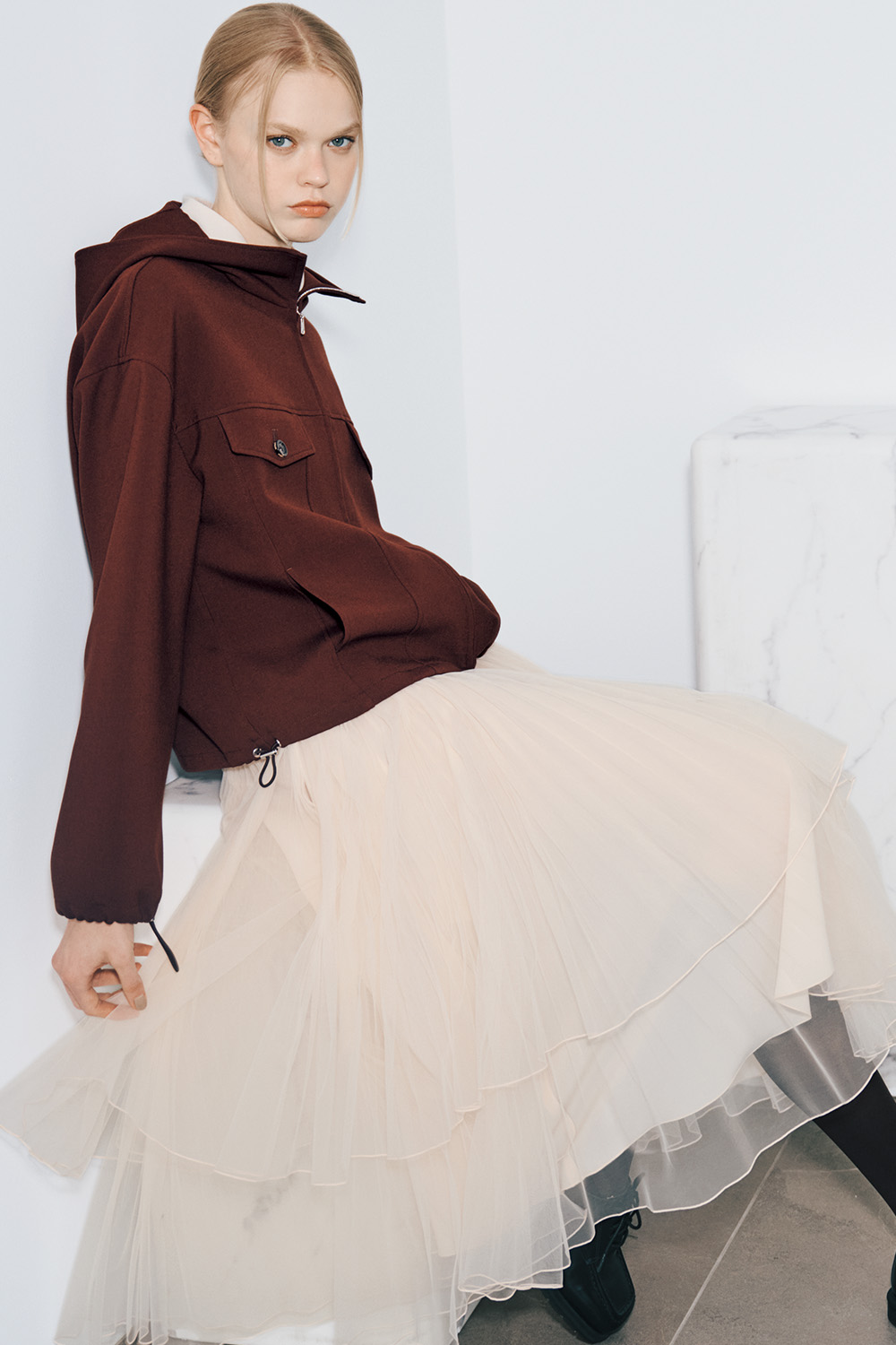 The model wears a cropped brown hooded jacket over a white collared shirt, a cream tiered tulle skirt, black tights, and black shoes.