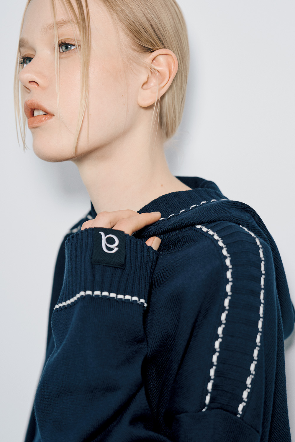 The model wears a navy knit hoodie with white contrast stitching along the hood and sleeves.