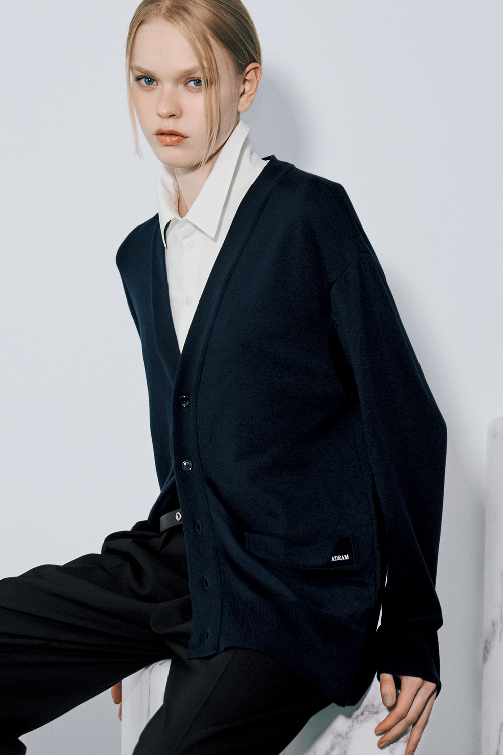 The model wears a navy V-neck cardigan with a patch pocket over a white collared shirt, a black belt, and black trousers.