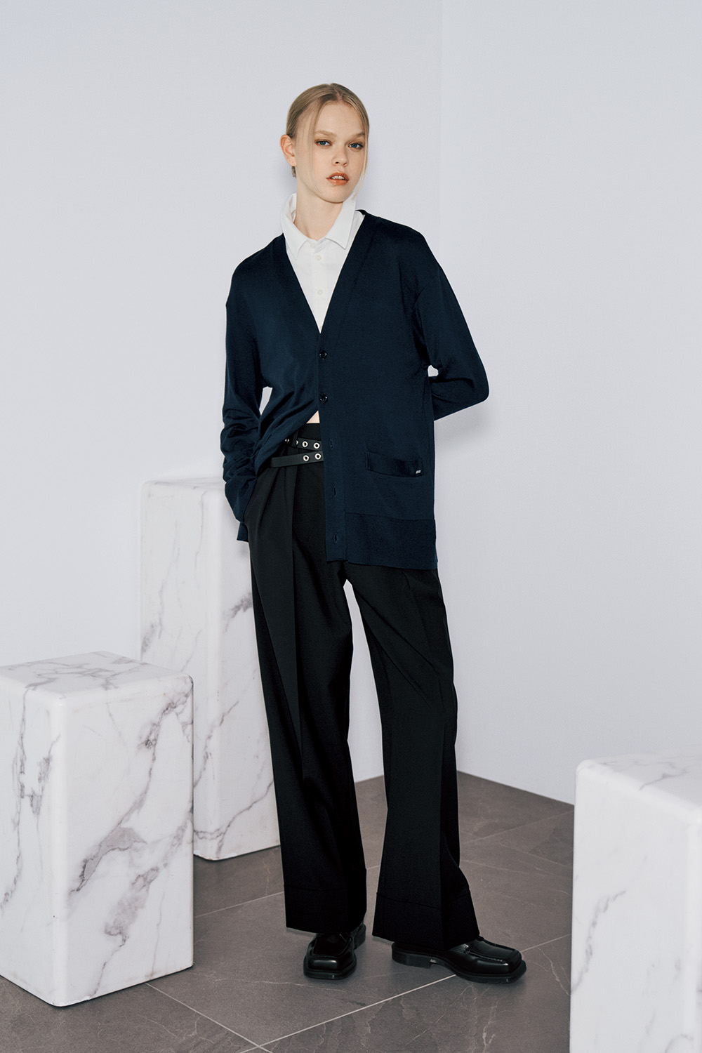 The model wears a navy V-neck cardigan over a white collared shirt, a black belt with silver eyelets, black wide-leg trousers, and black leather loafers.