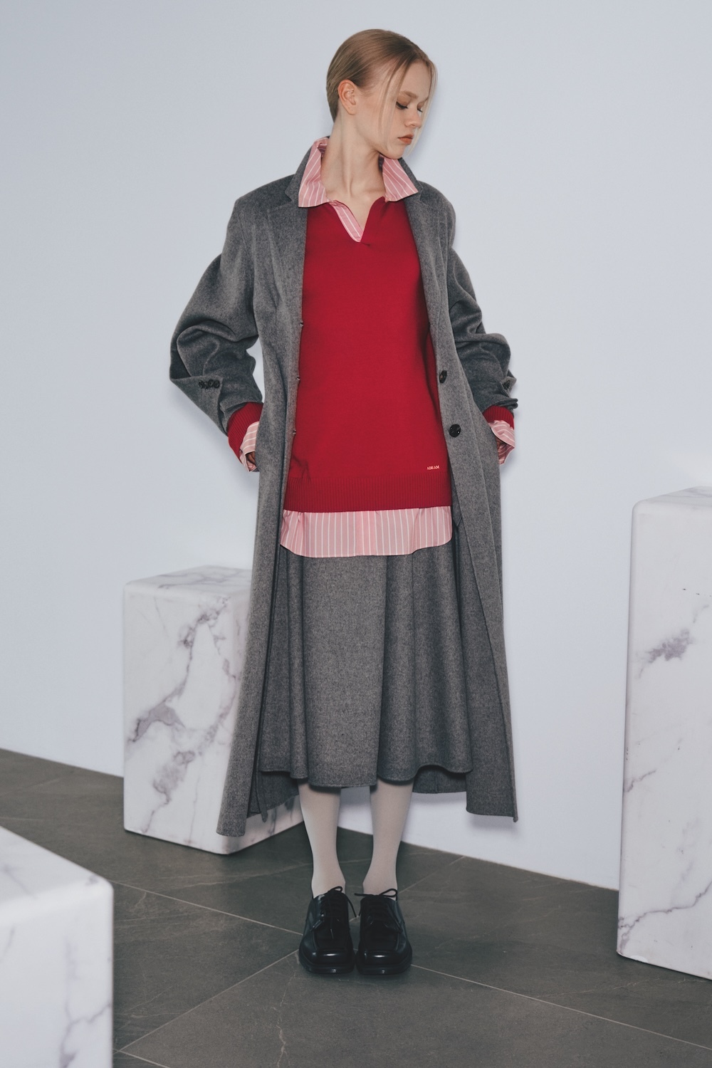 The model wears a long grey wool coat over a red knit polo sweater layered on a pink-and-white striped shirt, with a grey midi skirt, white tights, and black leather shoes.
