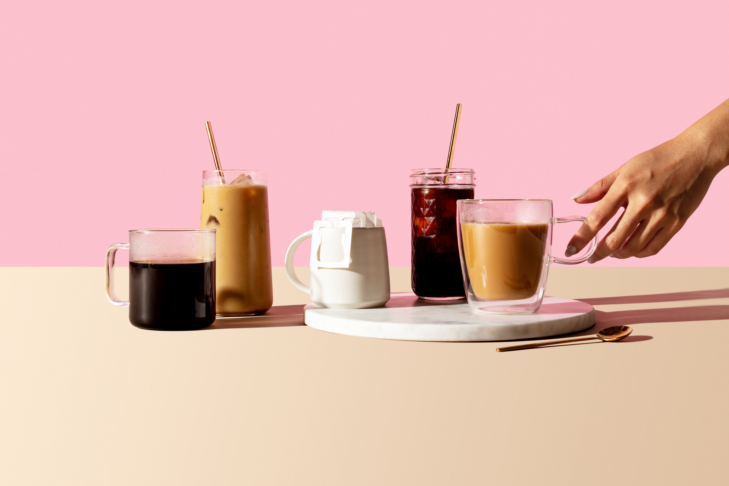 Clear glass mug with black coffee next to tall glass with iced latte and straw next to brewing coffee in white ceramic mug, next to iced black coffee with straw, next to latte in clear glass mug being picked up by a hand. 
