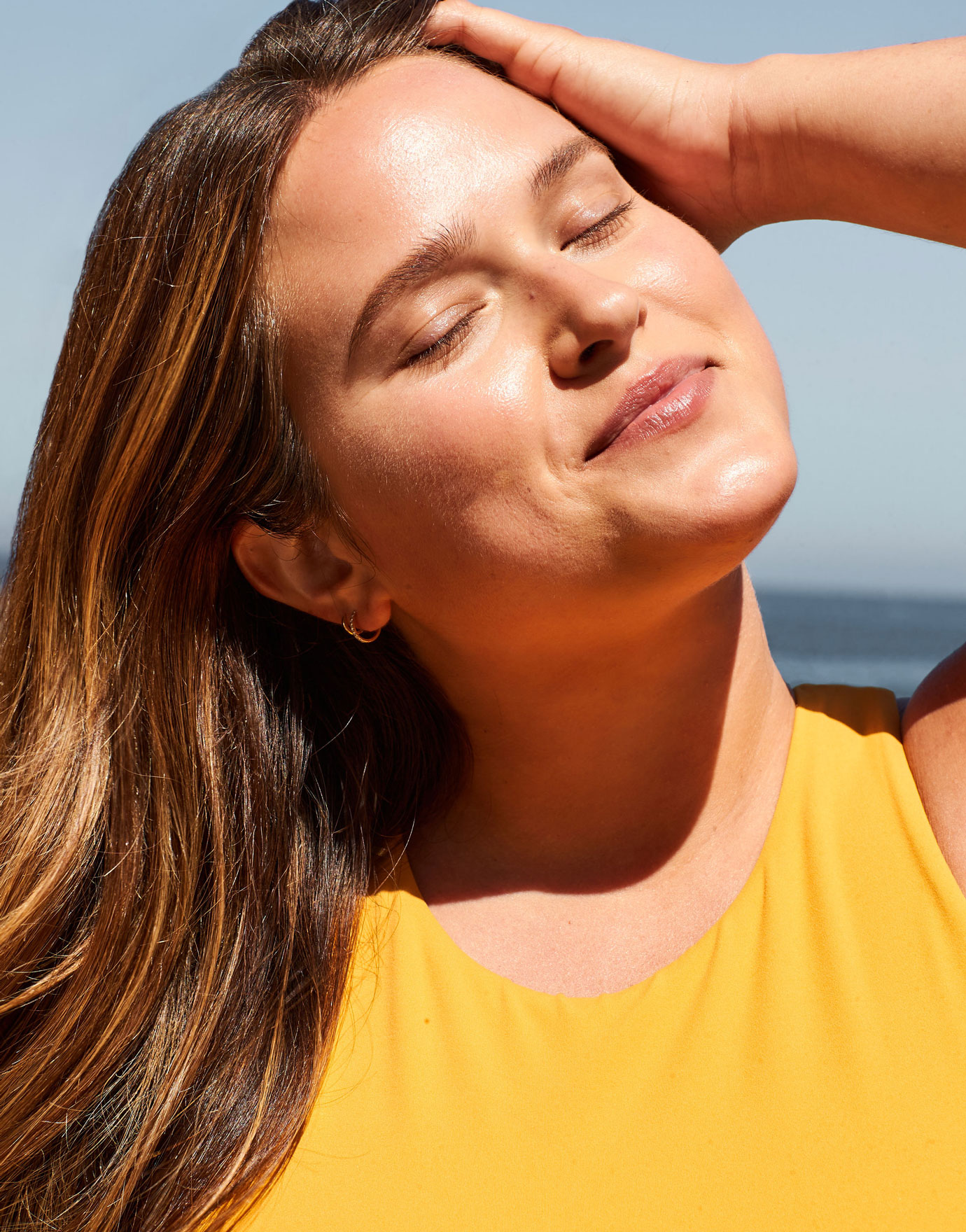 Woman with brown hair, a yellow top, and dewy, protected skin smiles serenely in the sunlight. 