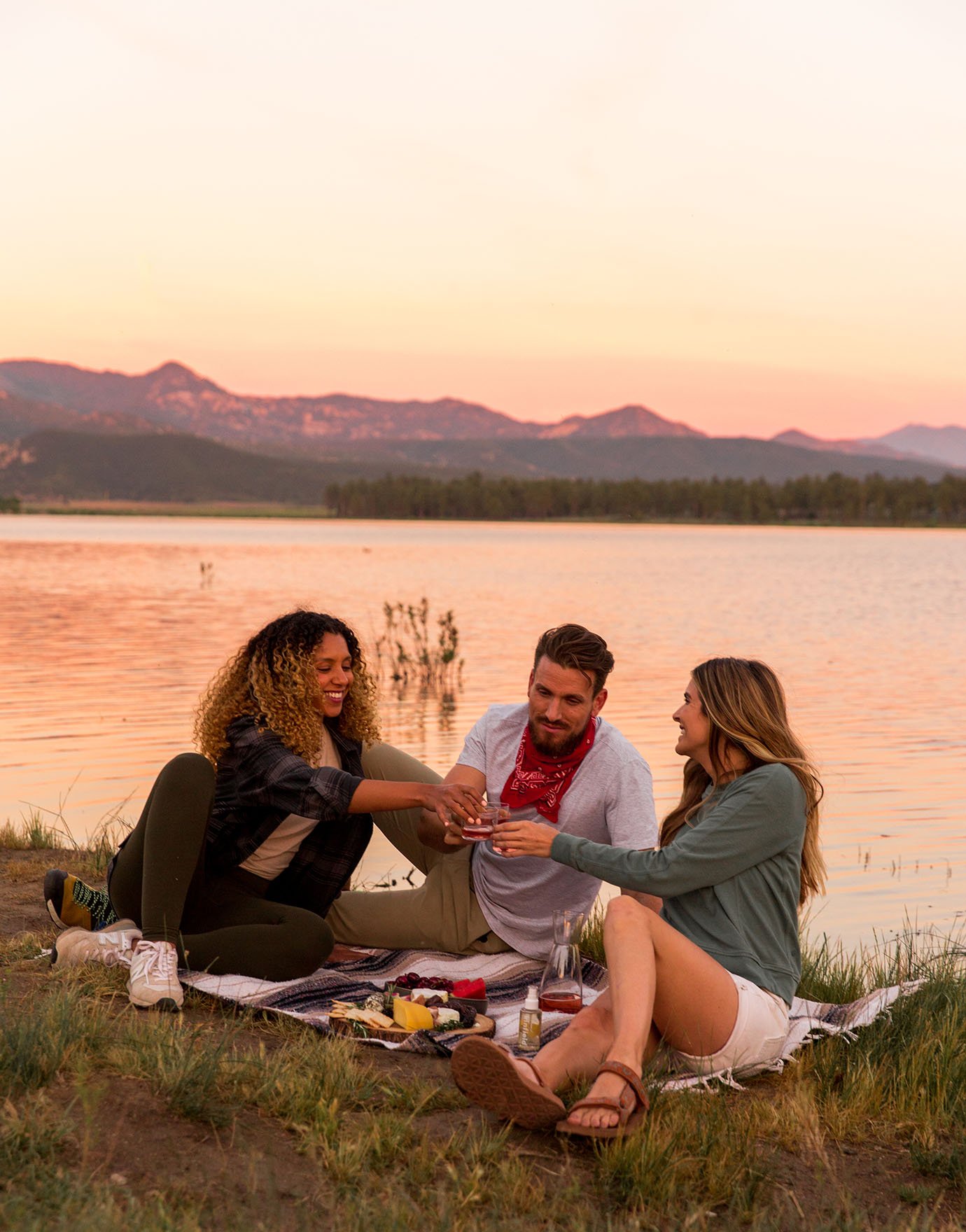 A group of two women and one man sit on a picnic blanket next to a lake while the sun sets.
