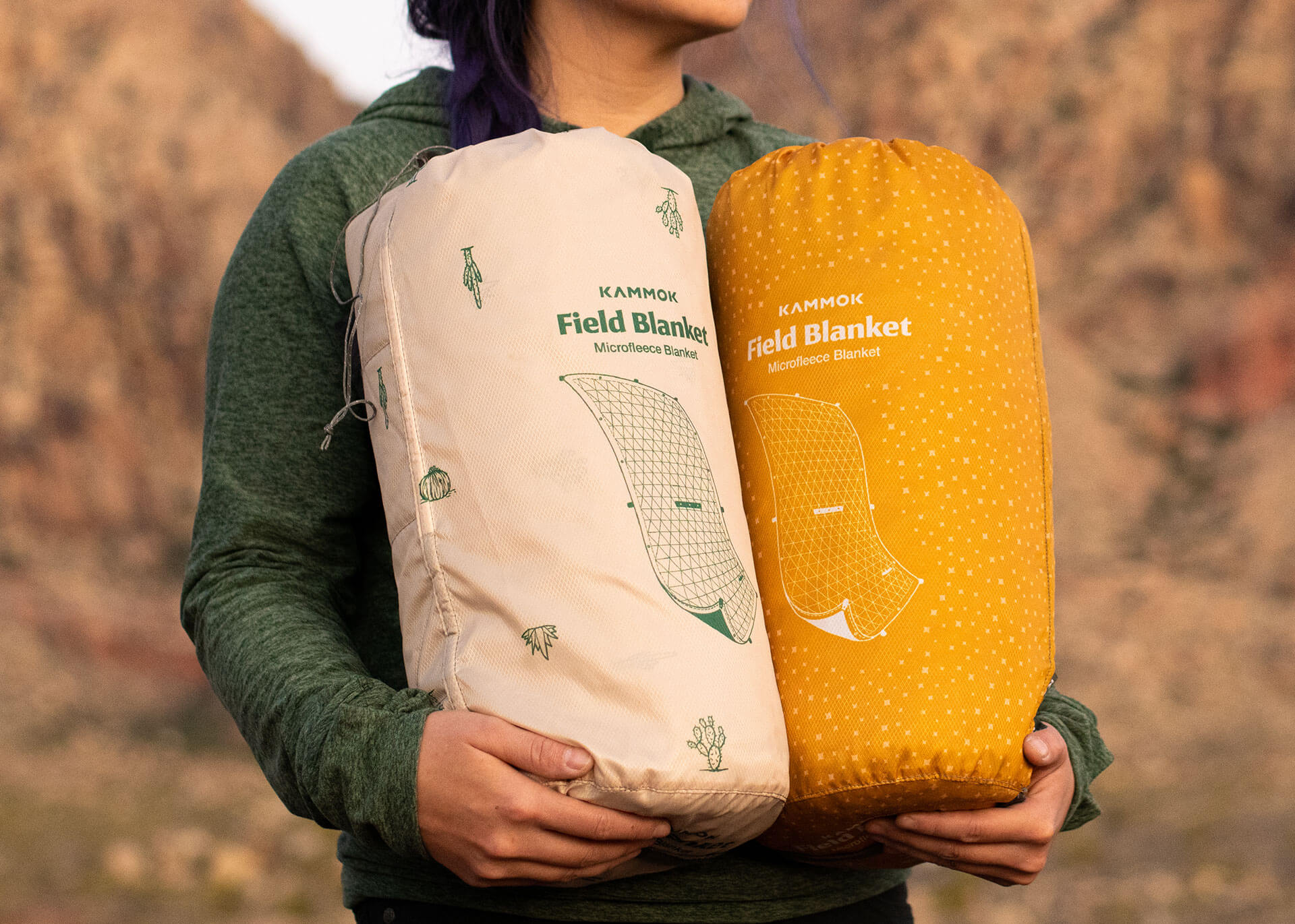 Woman holding two stuff sacks containing field blankets printed in front of a mountain backdrop.