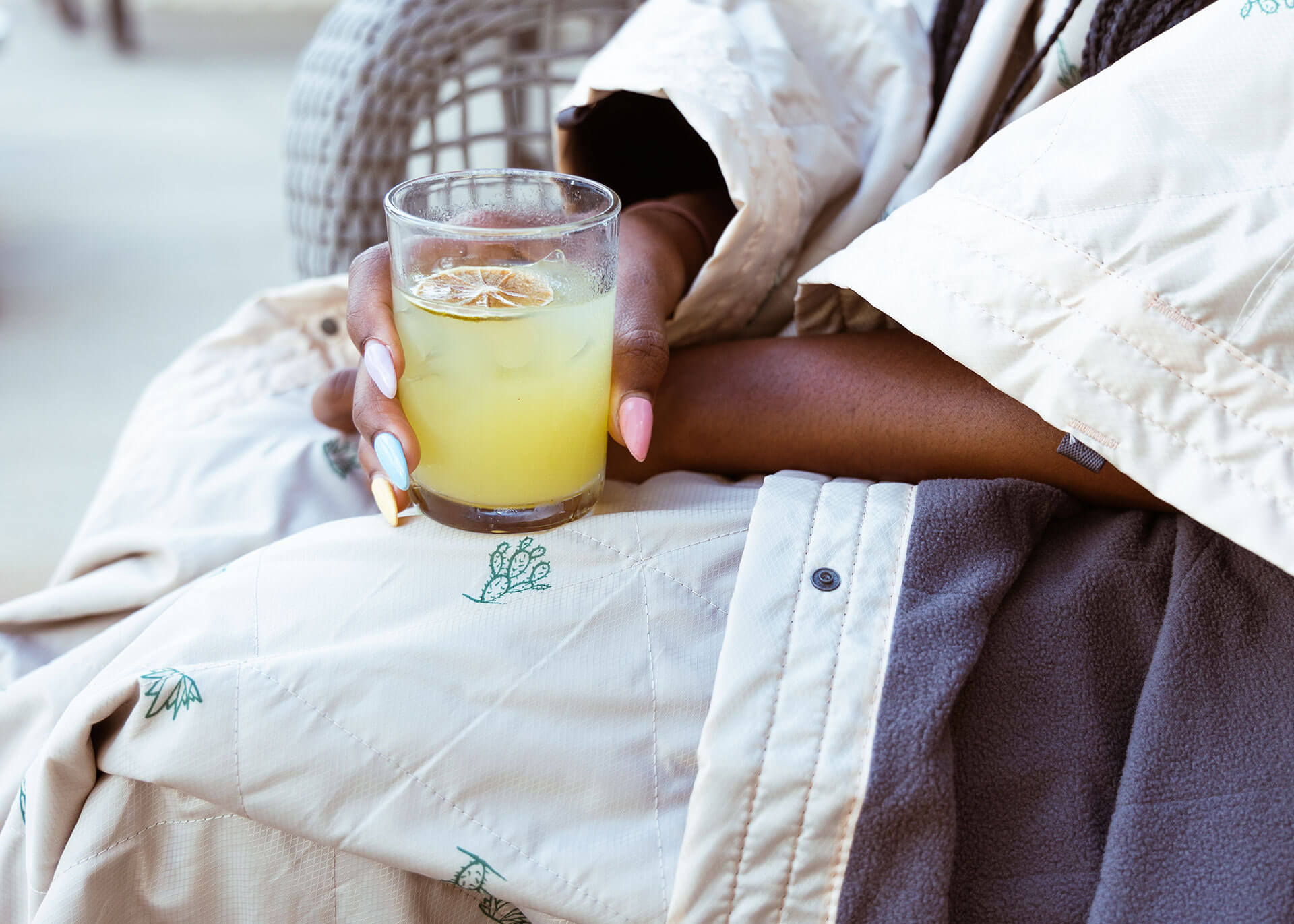 Woman holding a yellow drink in glass while wearing a Kammok Field Blanket Printed in sand tan pink color with cacti print. 