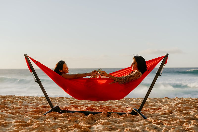 Two women sitting in a Kammok Roo Double two person hammock and swiflet portable hammock stand.