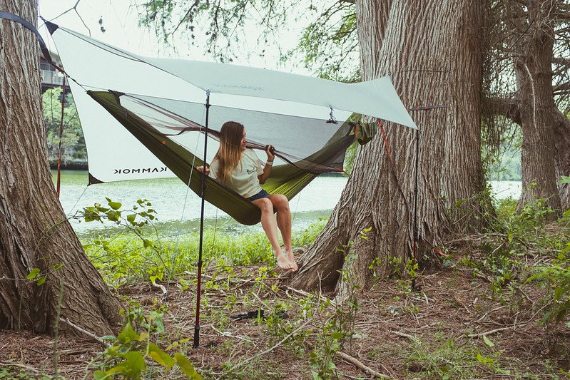 Woman sitting in Kammok Mantis all-in-one hammock tent with with built-in rainfly hung above in awning mode with hiking poles.