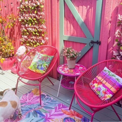 A polyester outdoor rug with a psychedelic exotic floral design, laid on a wooden deck surface in a pink garden setting.