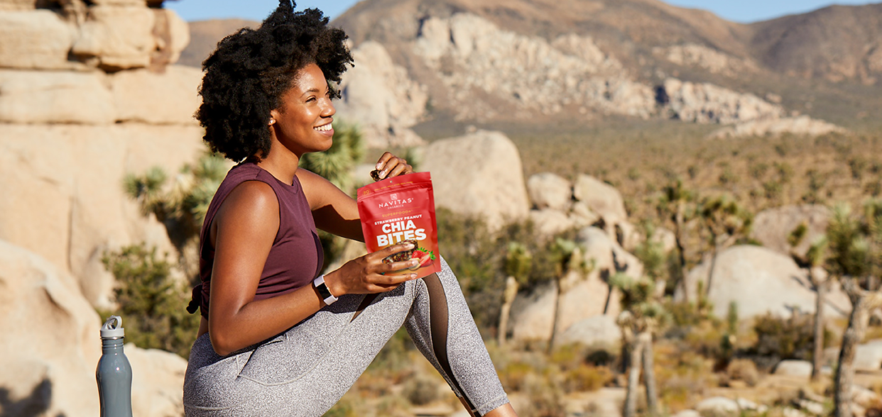 Person sitting on a rock outdoors, holding a bag of Navitas Organics Superfood+ Strawberry Peanut Chia BITES, smiling, with a scenic desert landscape in the background.