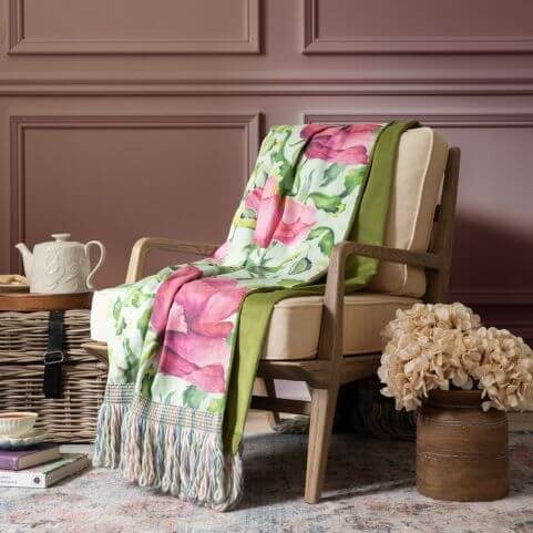 A sage fringed throw with a printed design of summer florals, draped over an arm chair alongside co-ordinating summer decor.