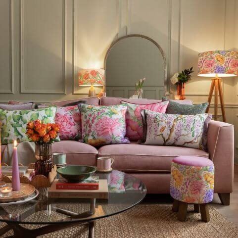 A pink living room sofa decorated with a series of vibrant floral cushions, alongside a matching footstool, lampshade and other spring and summer decor.