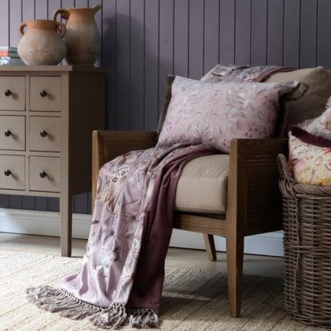 A fringed throw with a printed floral design in a luxurious violet shade, draped over a neutral arm chair with a matching floral cushion.