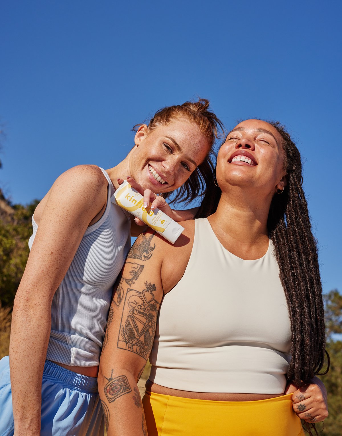 Two women stand together, smiling in the sun, while wearing athletic clothing and holding Cloud Cover Sunscreen.