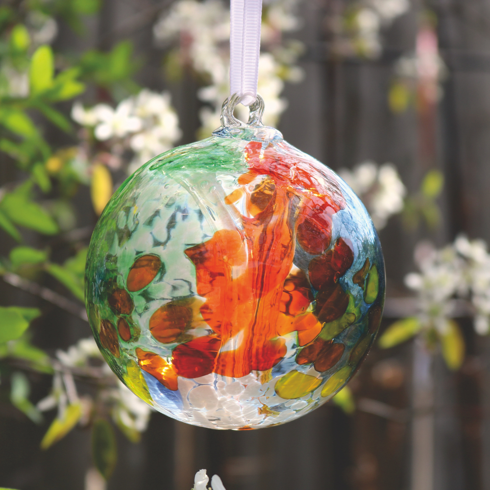 Glass orb with orange, white, yellow and green glass on the surface with white webbing pulled from bottom to top hanging on a white ribbon in front of greenery and white blooms.