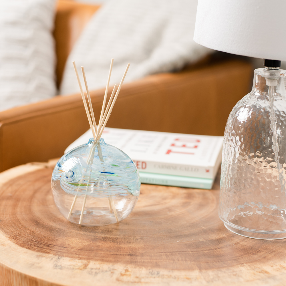 A clear glass orb with a flat bottom with white, blue and glass swirled on the top half. Five reeds are in a hole in the top of the orb. Displayed on a wooden table beside a lamp with a book and couch in the background.