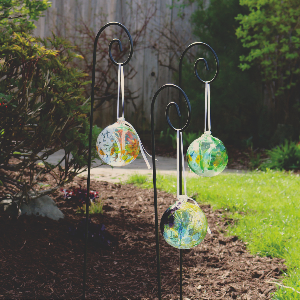 Three assorted Fairy Orbs hanging from white ribbons on black shepherd's hook stakes in a garden.