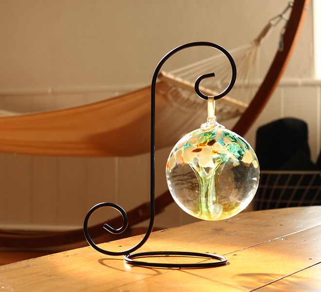 Clear glass orb with lime glass pulled through the middle and yellow, green, gold glitter and blue glass bits on top on a black ornament holder. Sunlight is shining in and there is a hammock in the background. 