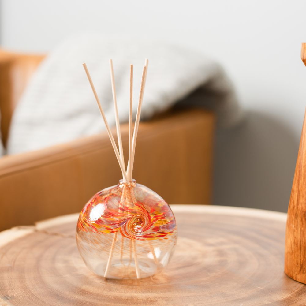 A clear glass orb with a flat bottom with red, orange and yellow glass swirled on the top half. Five reeds are in a hole in the top of the orb. Displayed on a wooden table.