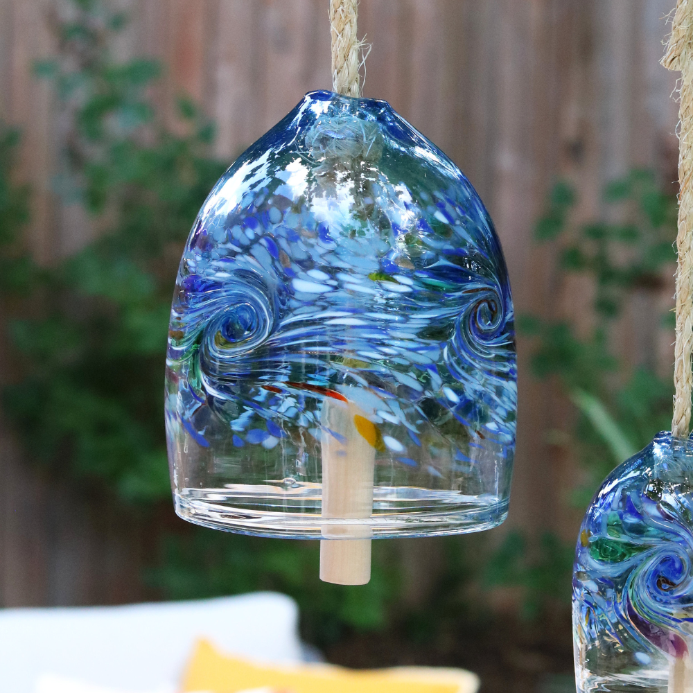 Two clear glass bells with blue, white red, green, yellow, opaque white and purple swirled across the middle. A wooden toggle and rope are pulled through the middle. Hanging amongst greenery in front of a wood fence.