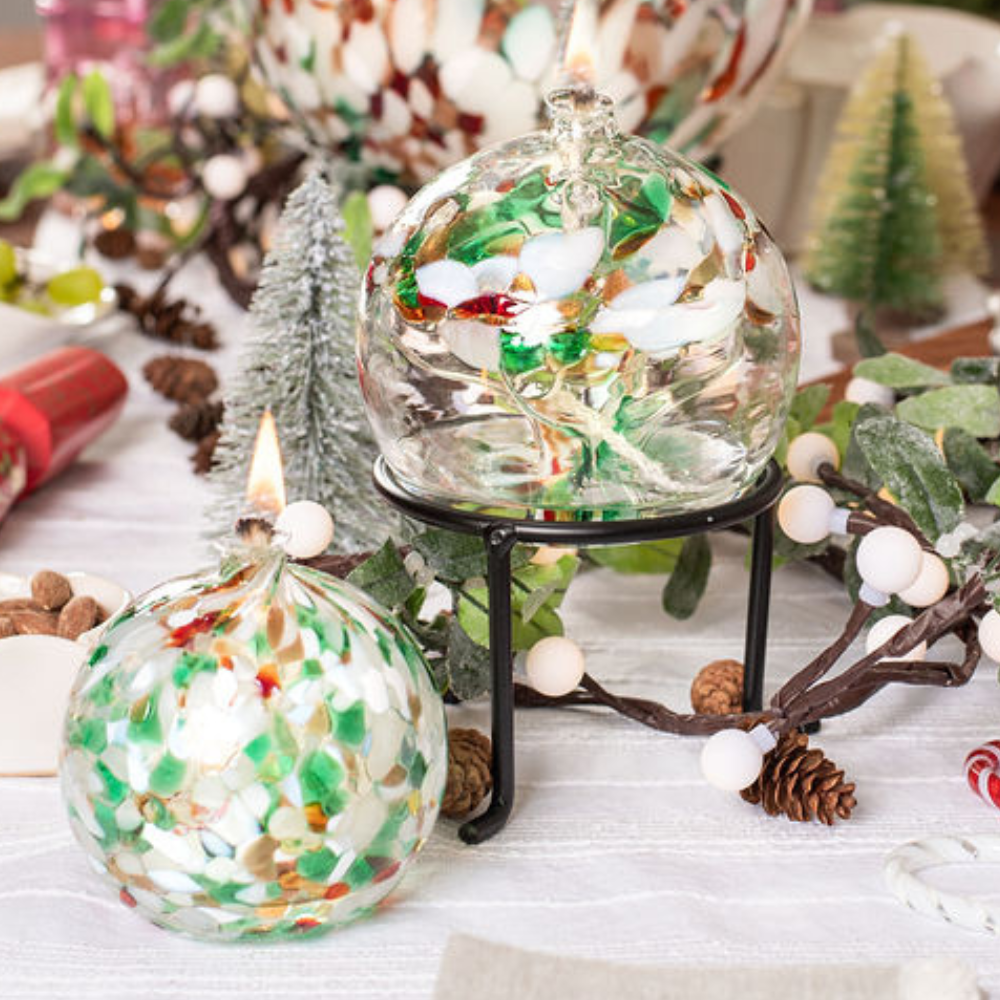 Lit clear glass flat bottom orb with red, green and clear glass pulled through the middle and white, red, green and gold glitter bits on top on a black three leg stand and lit flat bottom orb with white, red, green and gold glitter bits on the entire surface on a white table cloth amidst a holiday table setting.