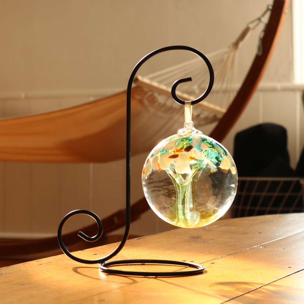 Clear glass orb with lime glass pulled through the middle and yellow, green, gold glitter and blue glass bits on top on a black ornament holder. Sunlight is shining in and there is a hammock in the background. 