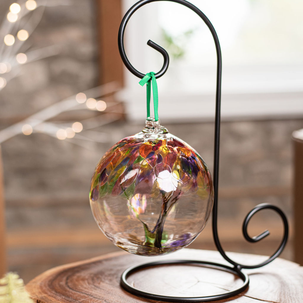 Clear glass orb with purple glass pulled through the middle and Yellow, Amber, Fuchsia, Purple, Brown and Green bits on top hanging from a green ribbon on a black ornament holder. Displayed in front of a window. 