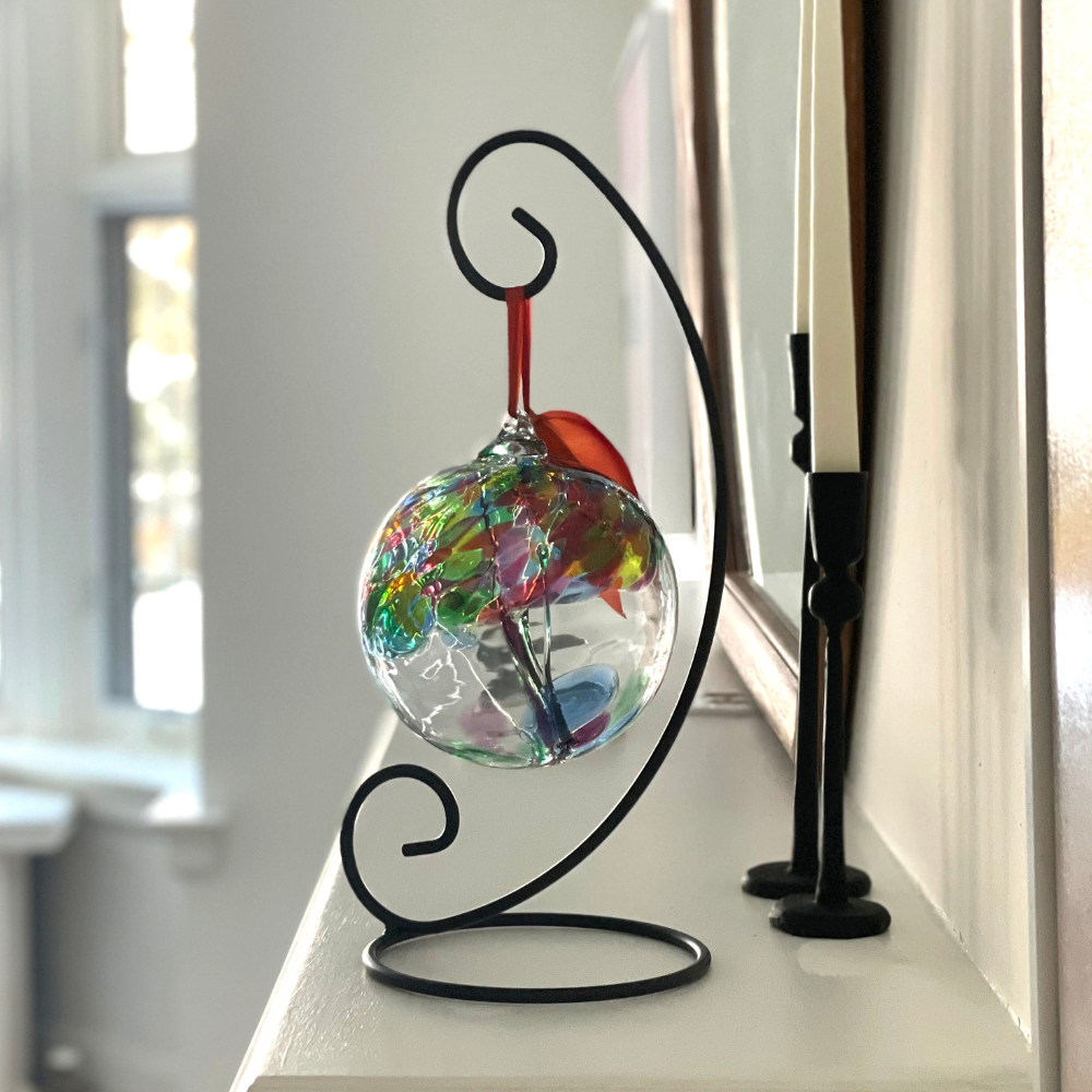 Clear glass orb with green glass pulled through the middle and red, green, lime, blue, teal and pink glass bits on top on a black curved ornament stand displayed on a white mantel. 