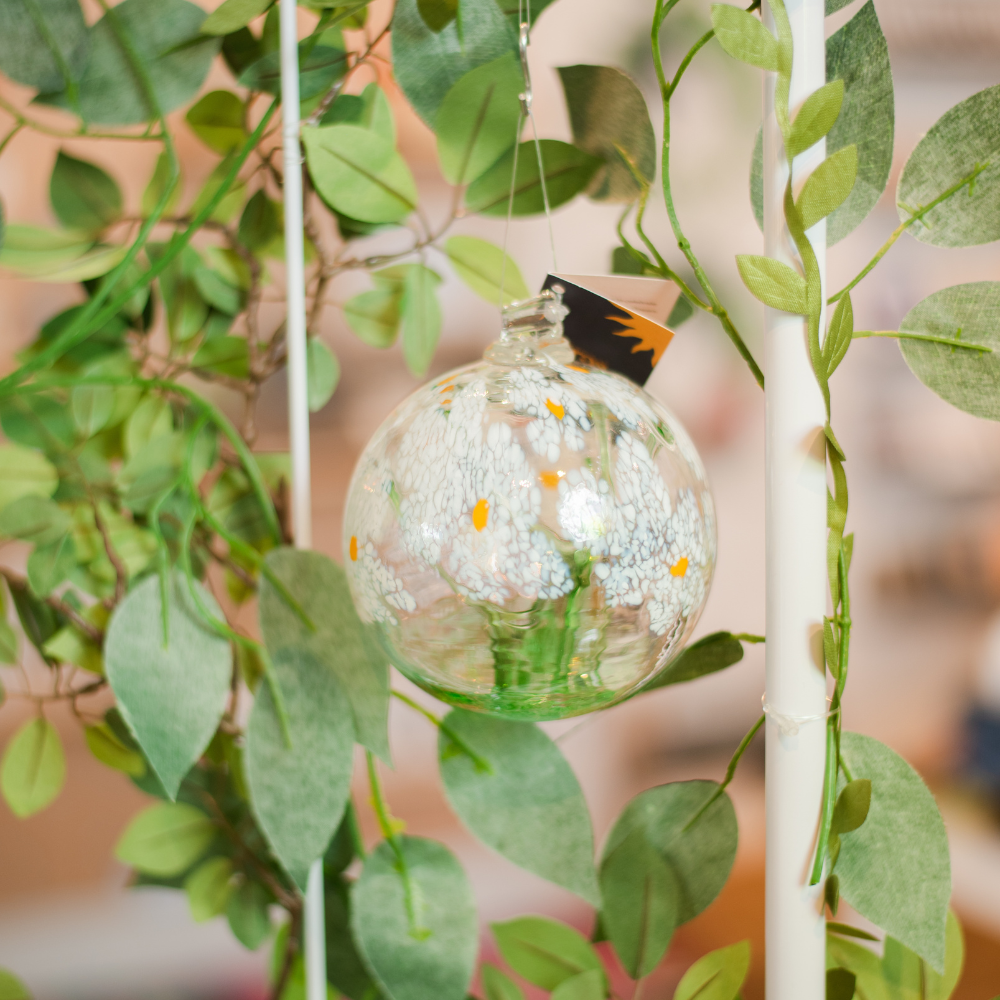 Clear glass orb with a green stem pulled from bottom to top to a canopy of white opaque glass blossoms and orange centres hanging amongst greenery. 