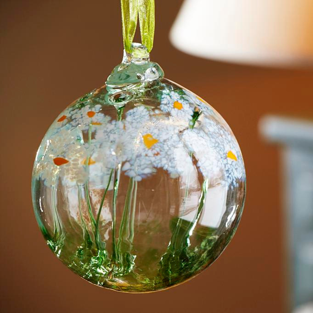 Clear glass orb with a green stem pulled from bottom to top to a canopy of white opaque glass blossoms and orange centres hanging in front of a red wall. 