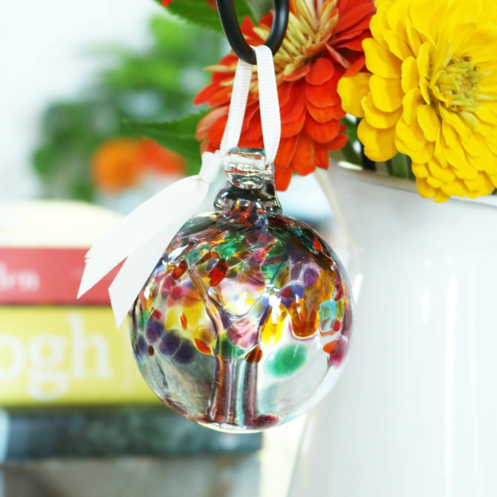 Clear glass orb with pink glass pulled through the middle and Purple, Pink, Orange, Green, Blue and Amber glass bits on top on a small black shepherd's hook stake in a vase with orange and yellow flowers. 