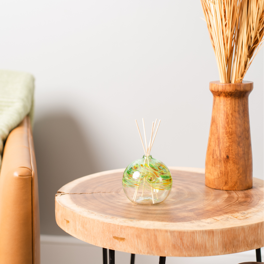 A clear glass orb with a flat bottom with green, amber, red and orange glass swirled on the top half. Five reeds are in a hole in the top of the orb. Displayed on a wooden table with a brown couch to the left and a brown vase to the right. 