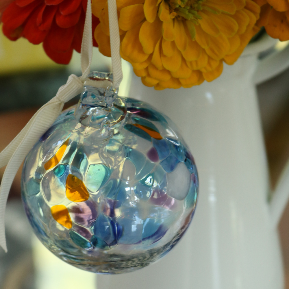 Clear glass orb with pink glass pulled through the middle and opaque white, blue, purple and orange bits hanging on a white ribbon with orange and red flowers in a white vase in the background