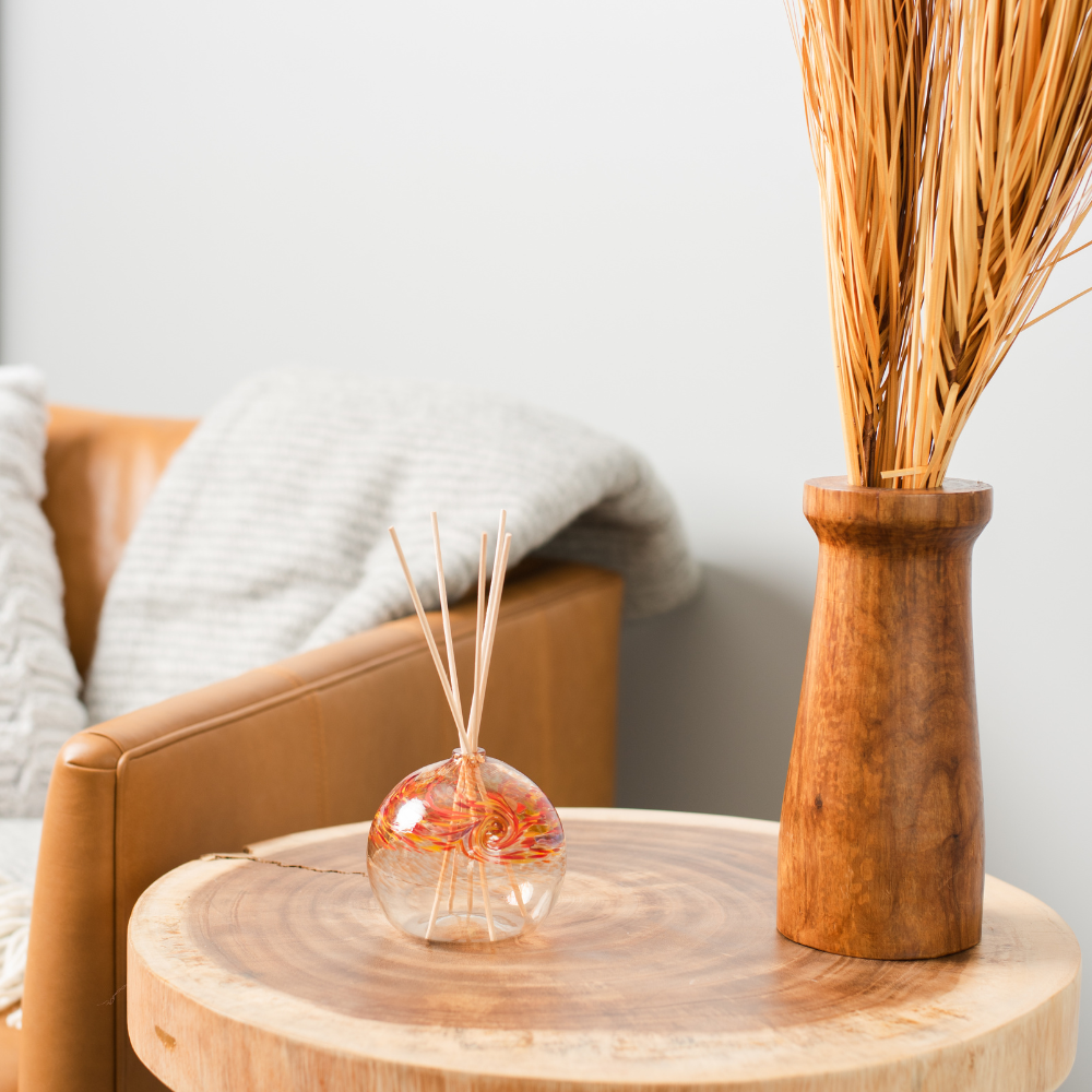 A clear glass orb with a flat bottom with red, orange and yellow glass swirled on the top half. Five reeds are in a hole in the top of the orb. Displayed on a wooden table with a brown leather couch in the background and a wood vase on the right.