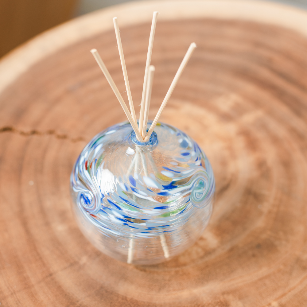 A clear glass orb with a flat bottom with blue, opaque white, red, green and purple swirled on the top half. Five reeds are in a hole in the top of the orb. Displayed on a wooden table. 