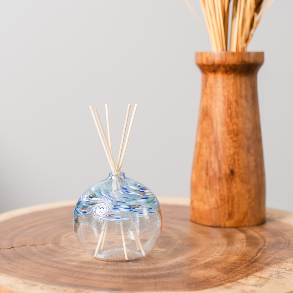 A clear glass orb with a flat bottom with blue, opaque white, red, green and purple swirled on the top half. Five reeds are in a hole in the top of the orb. Displayed on a wooden table. 