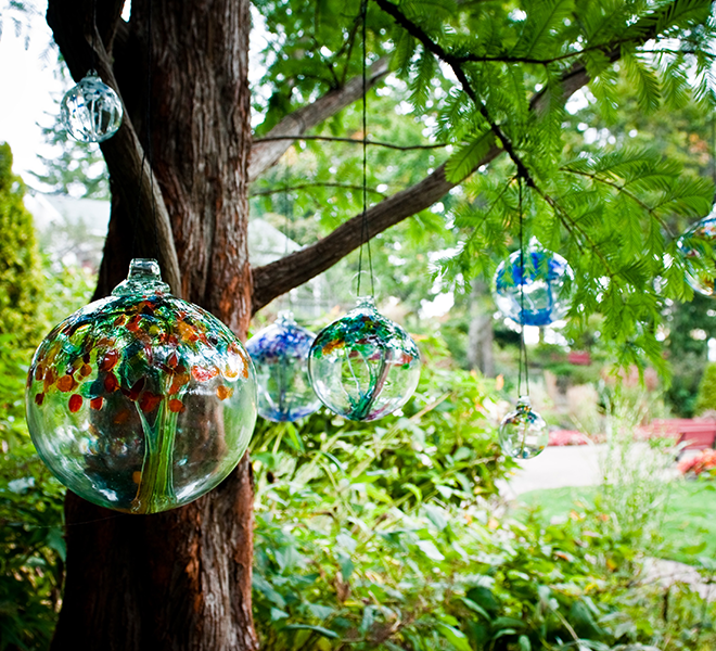 Clear glass orbs with all different coloured webbing, pulled from bottom to top, and glass bits on top of the orb. Hanging from black rope from brown tree branches with a green garden in the background. 