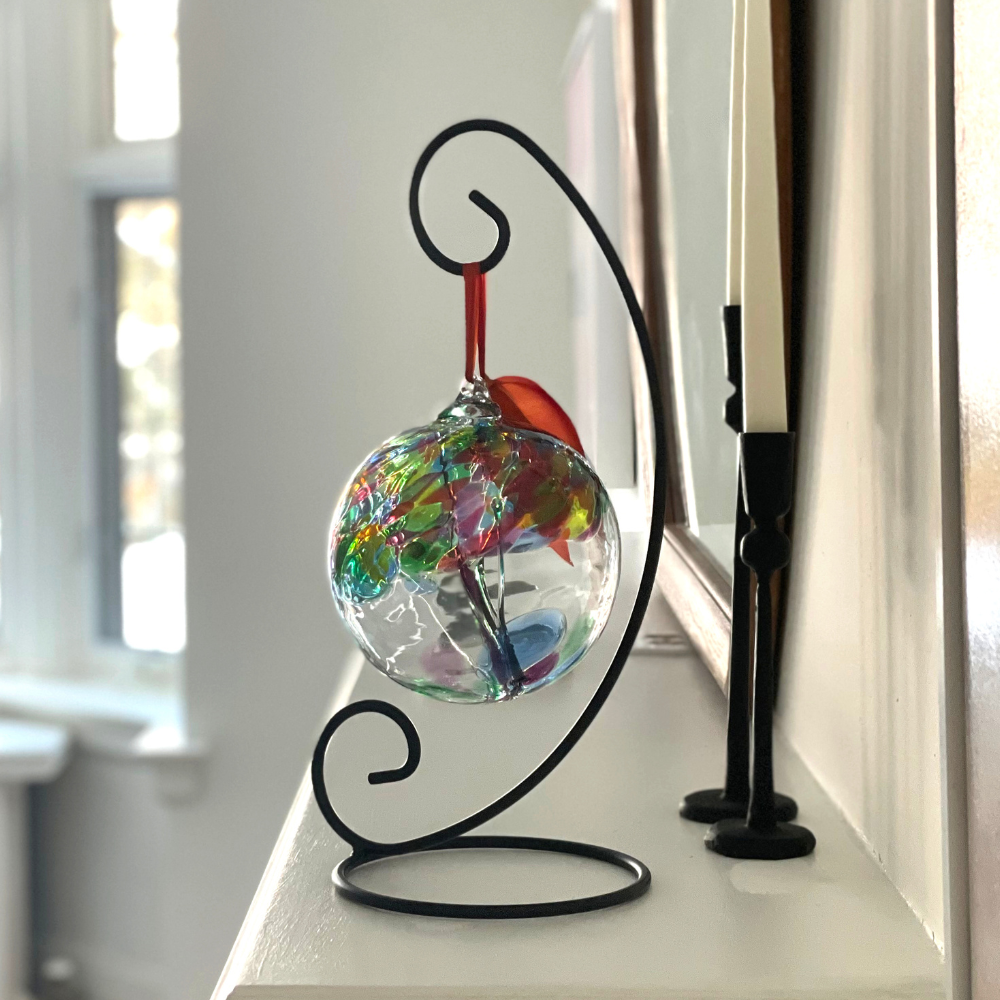 Clear glass orb with green glass pulled through the middle and red, green, lime, blue and pink glass bits on top on a black curved ornament stand displayed on a white mantel. 