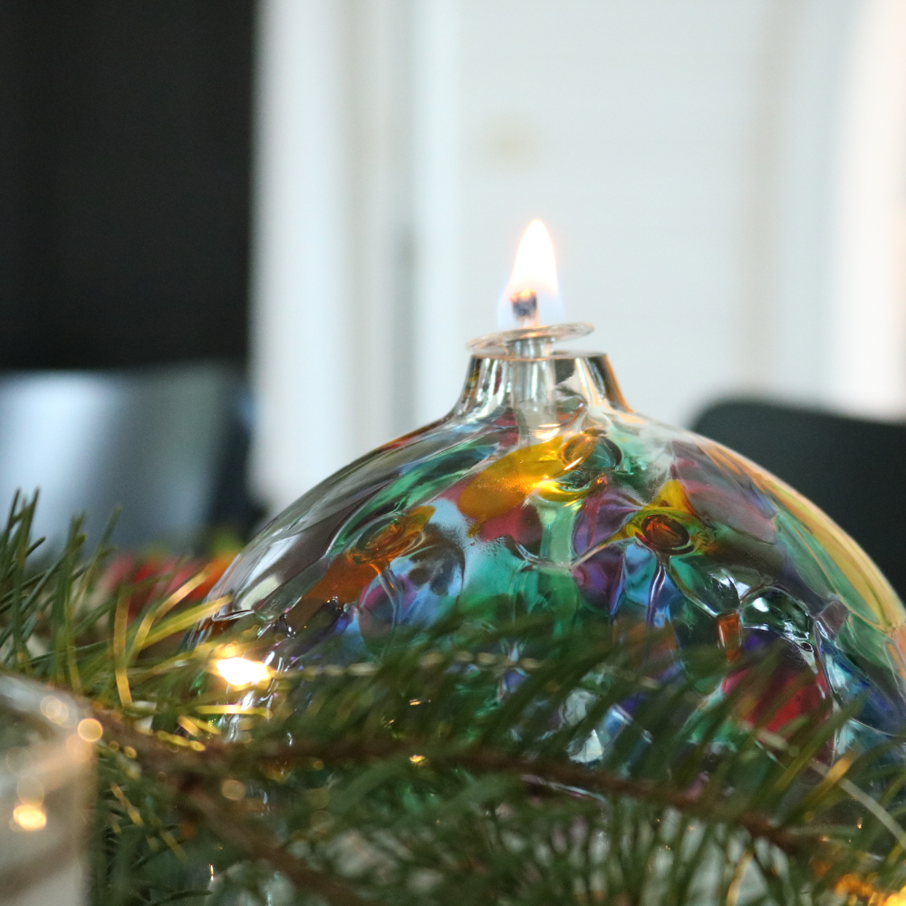 Lit clear glass flat-bottomed orb with a wick with pink glass pulled through the middle and Purple, Pink, Orange, Green, Blue and Amber glass bits on the top in the middle of a festive wreath. 