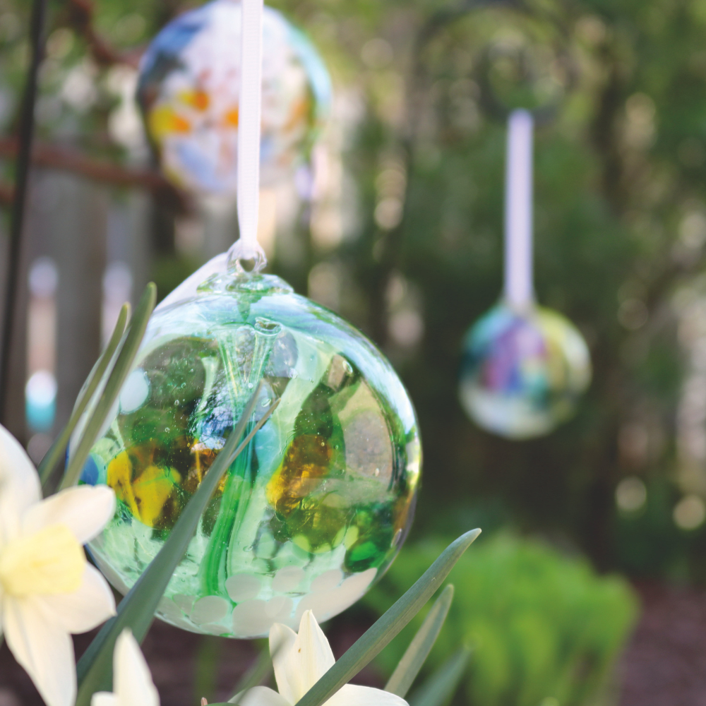 Glass orb with opaque white, green, orange, yellow and blue glass on the surface with white webbing pulled from bottom to top hanging on a white ribbon with glass orbs in the background in a garden.