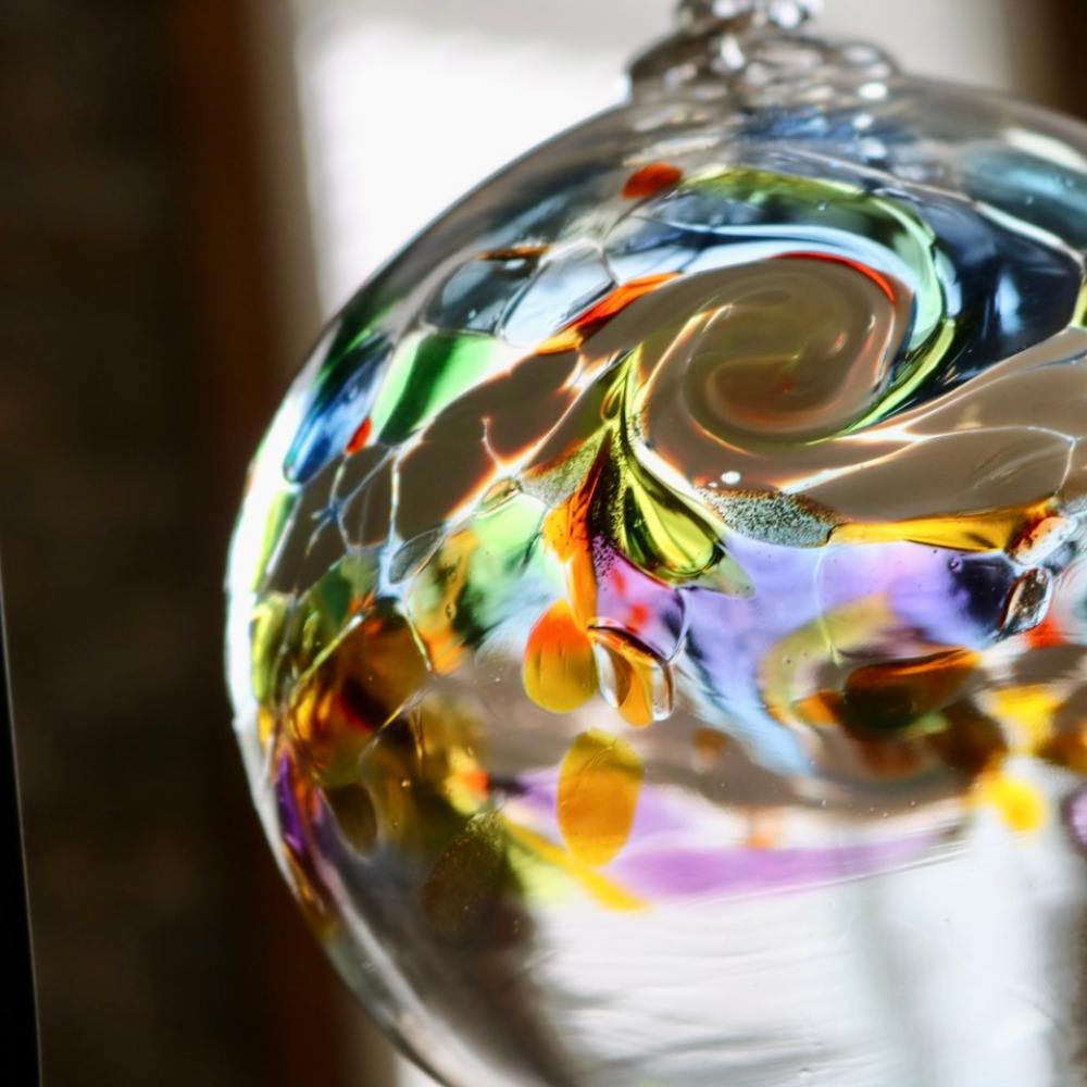 Glass orb with a colourful band across the middle. Blue, orange and green glass on top, white glass swirled in the middle and yellow, orange and red glass swirled below in front of a window