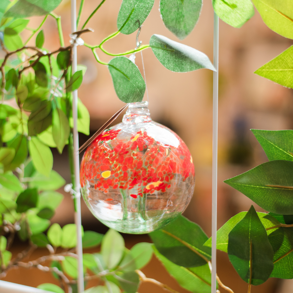 Clear glass orb with a green stem pulled from bottom to top to a canopy of red opaque glass blossoms and yellow centres hanging amongst greenery. 