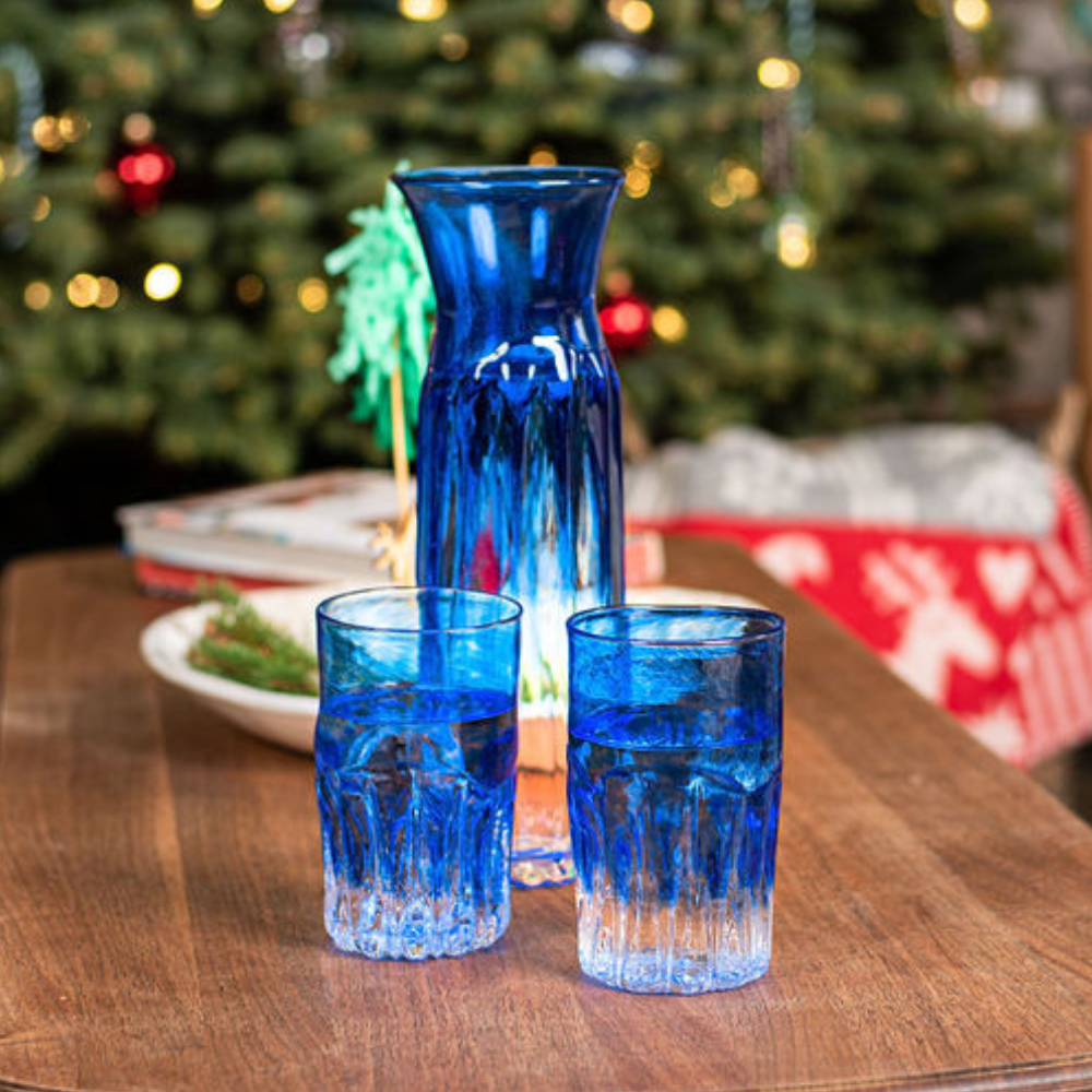 Carafe and two glasses with clear glass on the bottom and blue glass on the top. On a wood table with a Christmas Tree in the background.